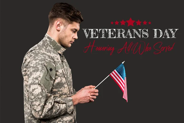 Sad man in military uniform holding american flag near veterans day lettering on grey — Stock Photo