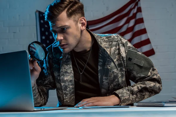 Handsome soldier holding magnifying glass near laptop in office — Stock Photo