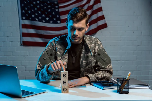 Schöner Militärmann, der neben Laptop sitzt und mit dem Finger auf Würfel mit SEO-Schriftzug zeigt — Stockfoto