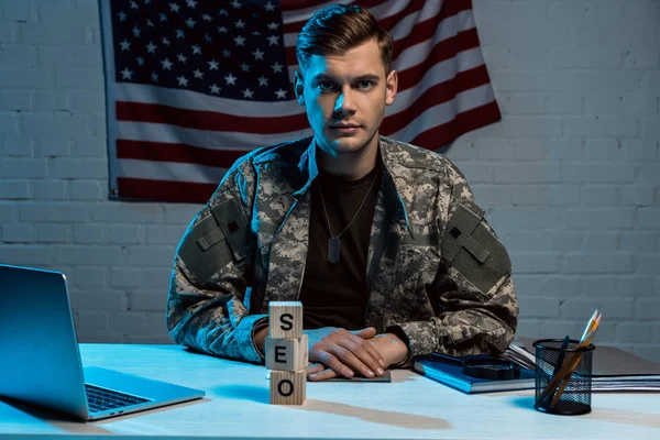 Guapo militar sentado con las manos apretadas cerca de la computadora portátil y cubos con letras seo — Stock Photo