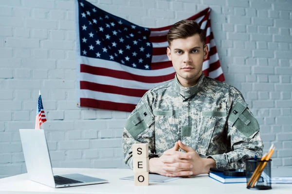 Handsome military man sitting with clenched hands near laptop and wooden cubes with seo lettering — Stock Photo