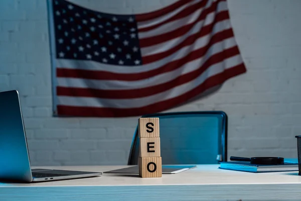 Würfel mit SEO-Schriftzug in der Nähe von Laptop und amerikanischer Flagge im modernen Büro — Stockfoto