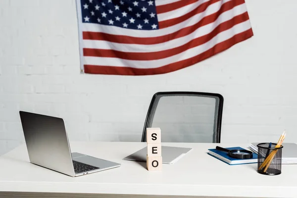 Wooden cubes with seo lettering near laptop and american flag in modern office — Stock Photo