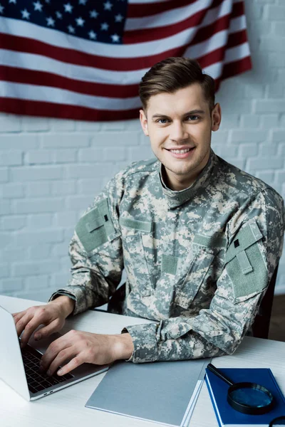 Cheerful soldier looking at camera while using laptop in office — Stock Photo