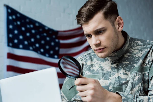 Low angle view of young soldier in uniform holding magnifying glass near laptop in office — Stock Photo