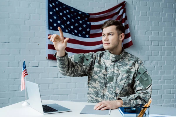 Bell'uomo in uniforme militare che punta con il dito e seduto vicino al portatile — Foto stock