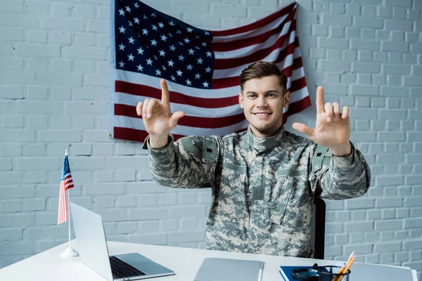 Hombre alegre en uniforme militar señalando con los dedos y sonriendo cerca del ordenador portátil - foto de stock