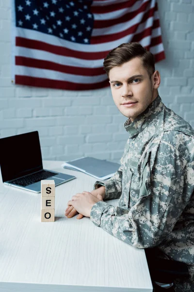 Handsome soldier looking at camera while sitting near laptop with blank screen and cubes with seo lettering — Stock Photo