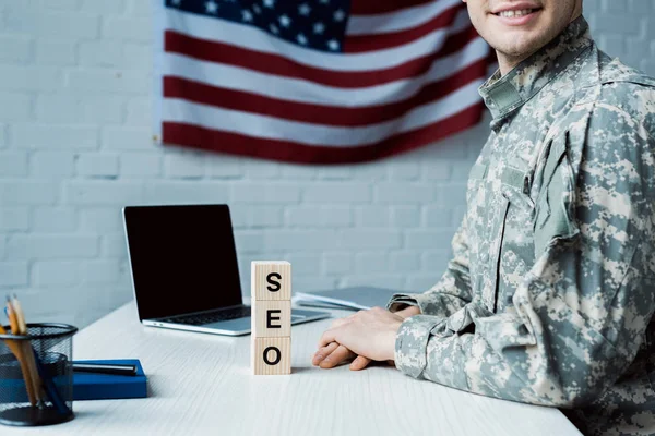 Vista cortada de soldado feliz perto de cubos de madeira com letras seo e laptop com tela em branco — Fotografia de Stock