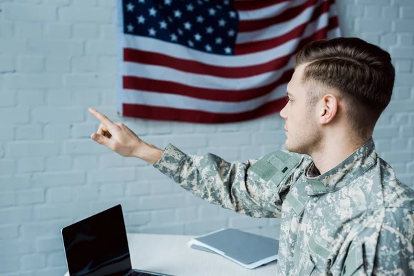 Guapo militar señalando con el dedo en la oficina cerca de la computadora portátil con pantalla en blanco - foto de stock