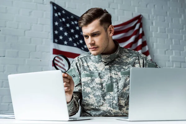 Guapo soldado sosteniendo lupa cerca de computadoras portátiles — Stock Photo