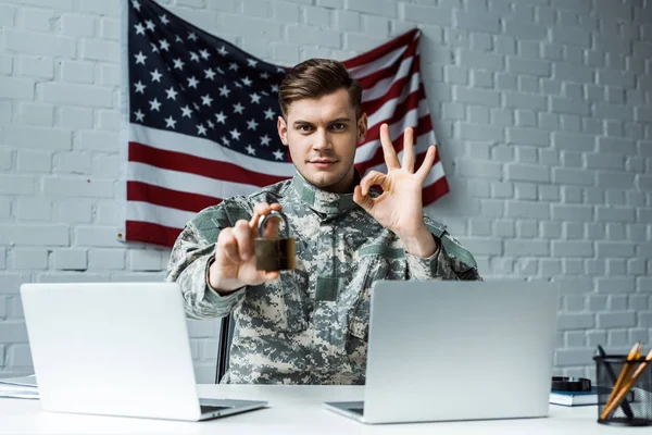 Hombre guapo en uniforme militar sosteniendo candado cerca de las computadoras portátiles y mostrando signo ok - foto de stock