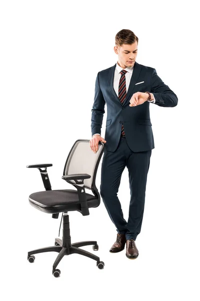 Selbstbewusster Geschäftsmann steht in der Nähe von Stuhl und schaut auf Uhr isoliert auf weiß — Stockfoto