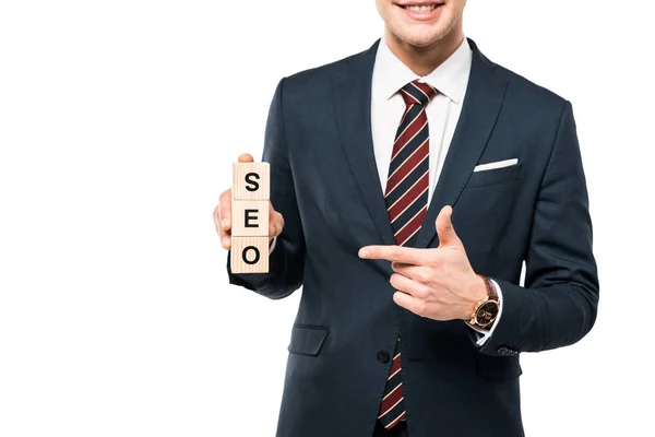 Vista cortada de empresário alegre apontando com o dedo em cubos de madeira com seo lettering isolado em branco — Fotografia de Stock