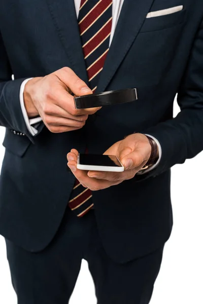 Vue recadrée de l'homme d'affaires en costume tenant loupe près du smartphone isolé sur blanc — Photo de stock