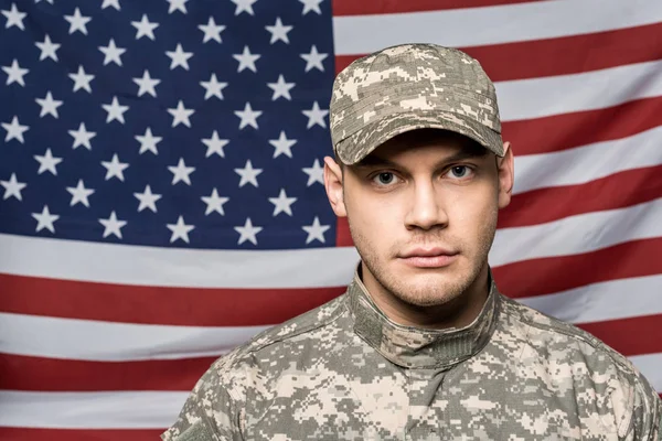 Handsome soldier in military uniform and cap looking at camera near flag of america — Stock Photo