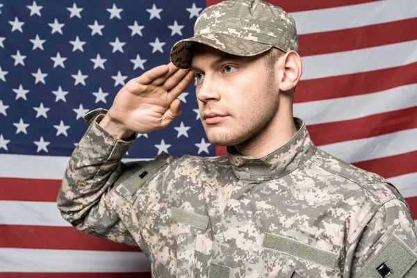 Guapo soldado en uniforme militar y gorra dando saludo cerca de la bandera de América - foto de stock