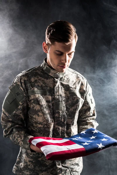 Hombre guapo en uniforme militar mirando la bandera americana en negro con humo - foto de stock