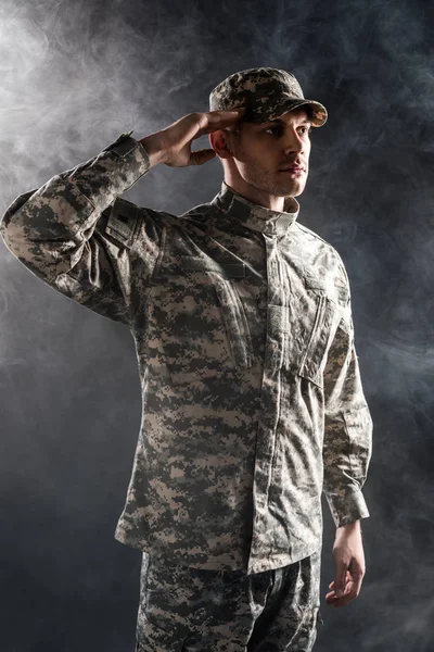 Soldier in military uniform and cap giving salute on black with smoke — Stock Photo
