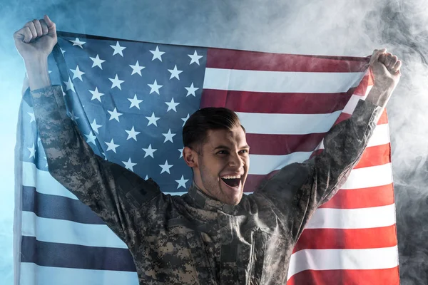 Soldado feliz gesticulando enquanto segurando bandeira americana em preto com fumaça — Fotografia de Stock