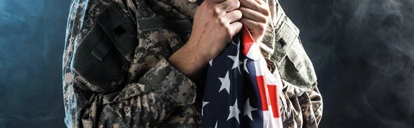 Plano panorámico de soldado sosteniendo bandera americana sobre negro con humo - foto de stock