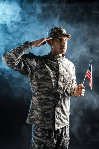 Soldado confiante em uniforme militar segurando bandeira americana e dando continência em preto com fumaça — Fotografia de Stock