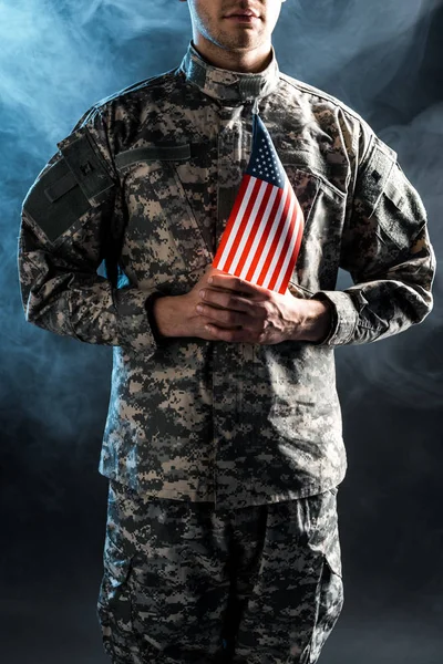 Обрізаний вид людини в камуфляжній формі, що тримає американський прапор на чорному з димом — стокове фото