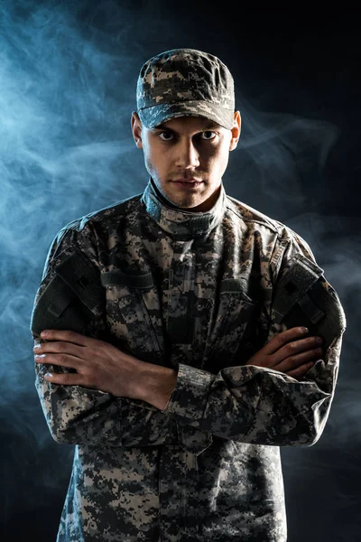 Confident soldier in military cap and uniform standing with crossed arms on black with smoke — Stock Photo