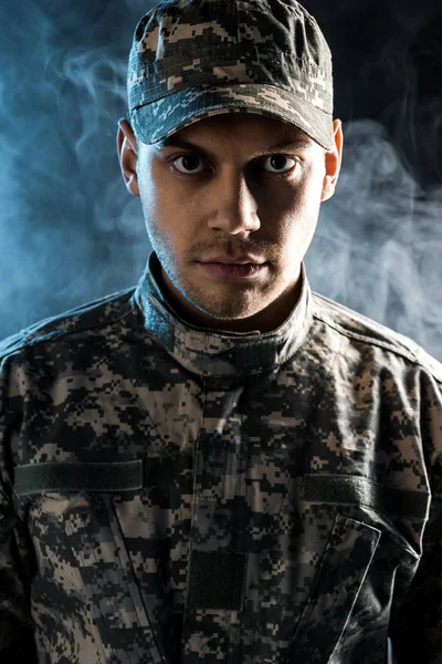 Confident soldier in military cap and uniform looking at camera on black with smoke — Stock Photo