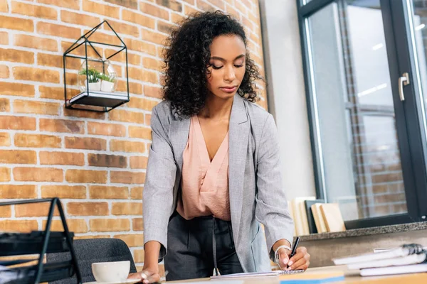 Attraktive afrikanisch-amerikanische Gelegenheitsunternehmerin schreibt in Notizbuch im Loft-Büro — Stockfoto