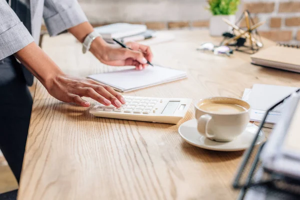 Vista recortada de la mujer de negocios ocasional afroamericana escribiendo en cuaderno y utilizando la calculadora en el escritorio - foto de stock