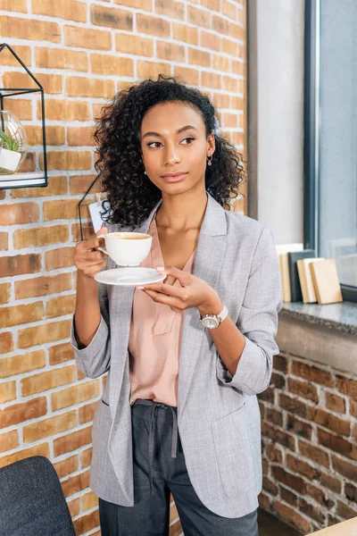 Hermosa afroamericana ocasional mujer de negocios sosteniendo taza de café con platillo en la oficina - foto de stock