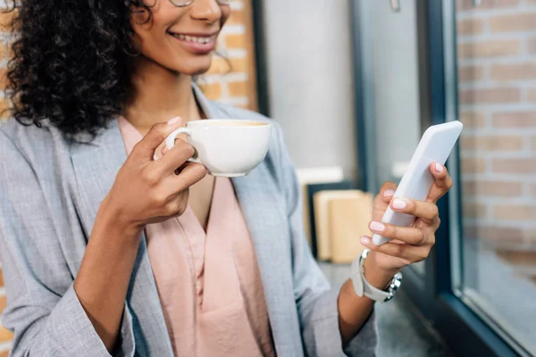 Vista recortada de la mujer de negocios ocasional afroamericana con taza de café usando teléfono inteligente - foto de stock