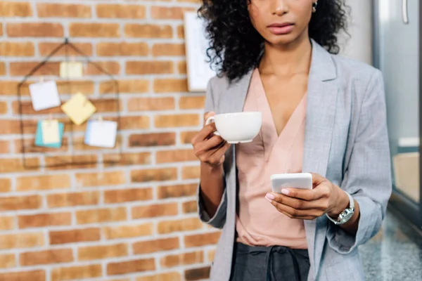 Vista recortada de la mujer de negocios ocasional afroamericana con taza de café usando teléfono inteligente - foto de stock