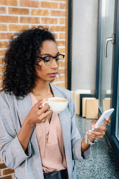 Ernsthafte afrikanisch-amerikanische Gelegenheitsunternehmerin in Gläsern mit Kaffeetasse mit Smartphone — Stockfoto