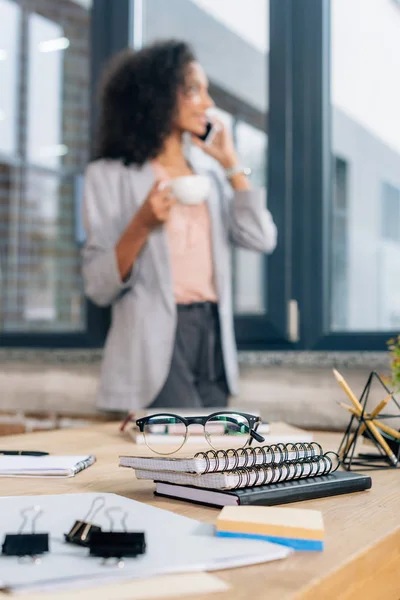 Enfoque selectivo de portátiles y gafas en el escritorio cerca de afroamericana Casual empresaria hablando en el teléfono inteligente - foto de stock