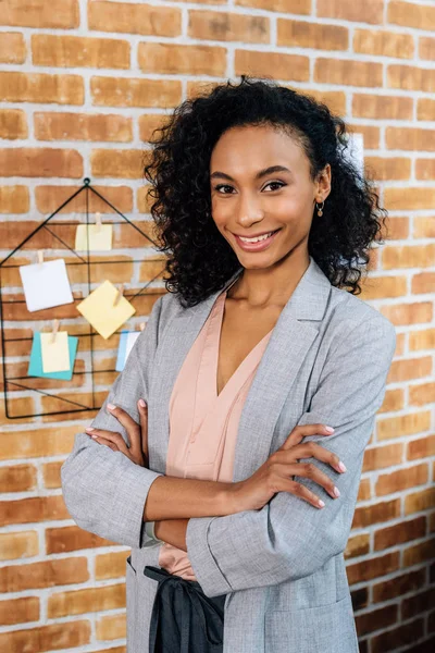 Sonriente afroamericana Casual mujer de negocios con los brazos cruzados en la oficina loft - foto de stock