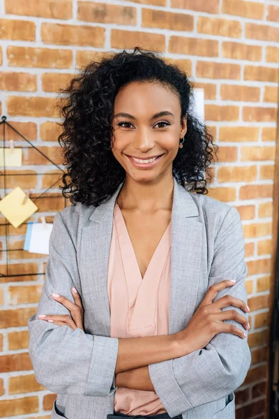 Sonriente afroamericana Casual mujer de negocios con los brazos cruzados en la oficina loft - foto de stock