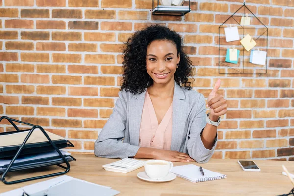 Sonriente ocasional afroamericana mujer de negocios mostrando el pulgar hacia arriba en escritorio de la oficina - foto de stock