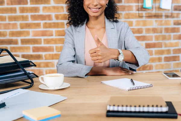 Обрезанный вид на улыбающуюся африканскую деловую женщину, показывающую большой палец на рабочем столе — стоковое фото