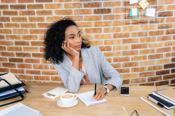 Afrikanisch-amerikanische Gelegenheitsunternehmerin sitzt mit Notizbuch und Stift am Schreibtisch im Büro — Stockfoto