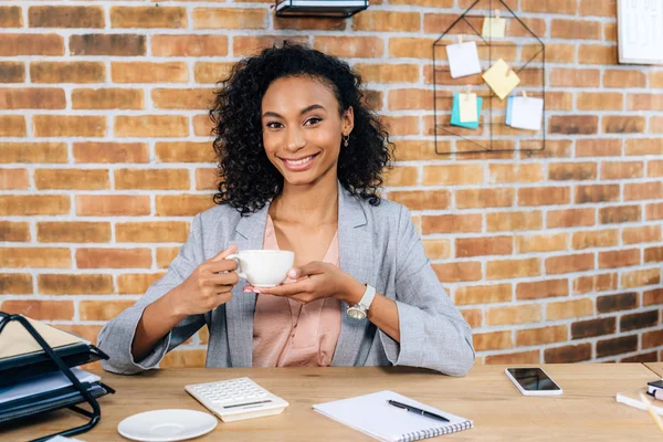 Sonriente afroamericana Casual mujer de negocios en el escritorio con taza de café en la oficina - foto de stock