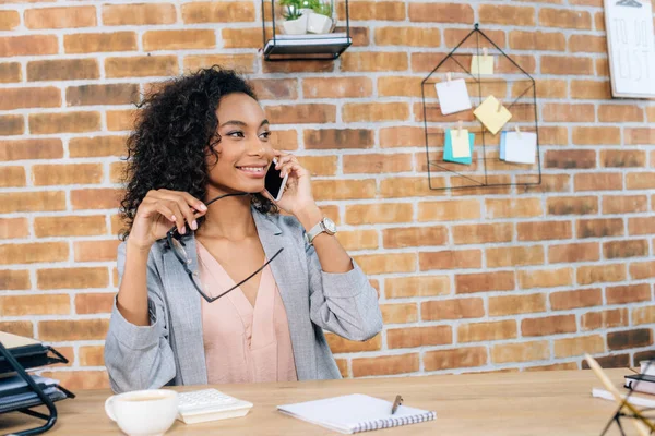 Lächelnde afrikanisch-amerikanische Gelegenheitsunternehmerin, die am Schreibtisch auf dem Smartphone spricht — Stockfoto