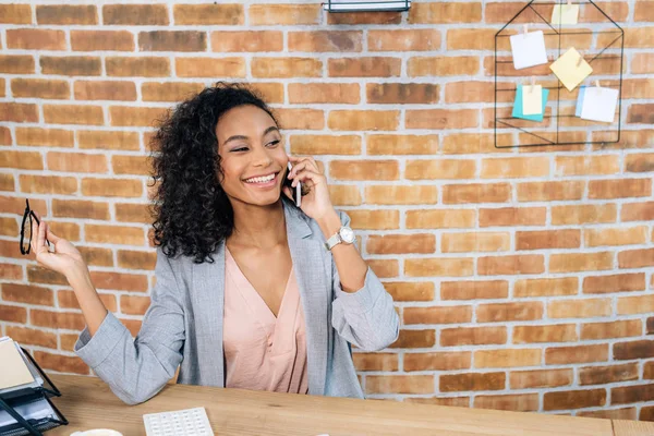 Усміхнена афроамериканка Випадкова бізнес-леді розмовляє на смартфоні за столом — стокове фото