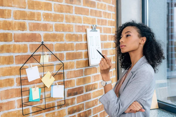 Lässige afrikanisch-amerikanische Geschäftsfrau mit Stift in der Nähe von Klemmbrett im Loft-Büro — Stockfoto