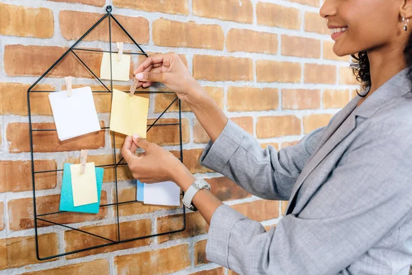 Vista recortada de la mujer de negocios afroamericana casual colgando notas adhesivas con alfileres en la oficina loft - foto de stock