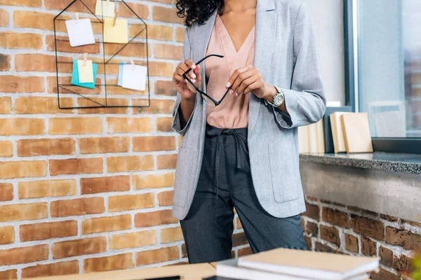 Vista ritagliata di casuale donna d'affari africana americana che tiene gli occhiali in ufficio loft — Foto stock