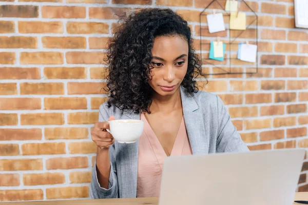 Africano americano Casual mujer de negocios con taza de café utilizando el ordenador portátil en la oficina - foto de stock