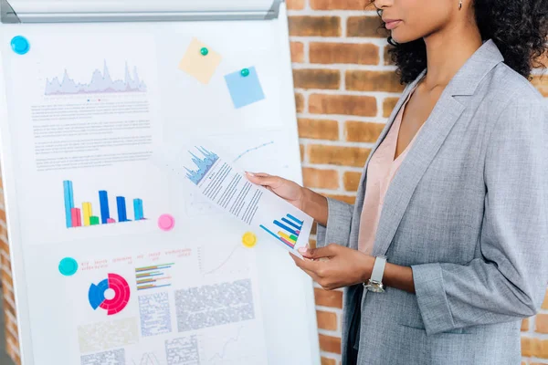 Vista recortada de la mujer de negocios ocasional afroamericana sosteniendo papel con gráfico cerca de flipchart en la oficina loft - foto de stock