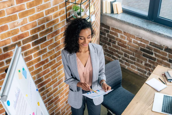 Hochwinkelaufnahme einer afrikanisch-amerikanischen Geschäftsfrau, die im Loft-Büro Papier in der Nähe von Flipchart hält — Stockfoto